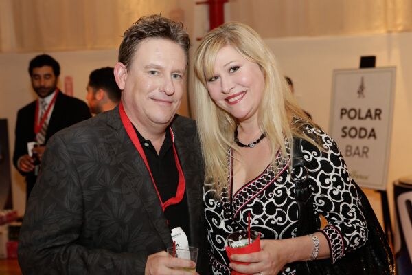 A man in a patterned dark jacket and a blonde woman in a black and white top smile at the camera, each holding a drink. A sign reading Polar Soda Bar stands behind them.