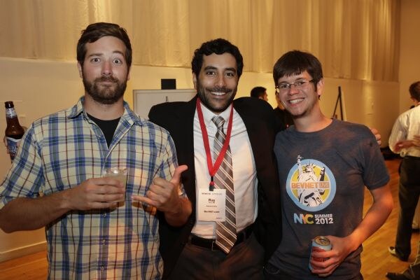 Ray of BevNET.com, wearing a suit, tie and red lanyard, smiles between two men. The man on the left holds a drink and gives a thumbs up; the man on the right wears a BevNET Live NYC 2012 T-shirt.