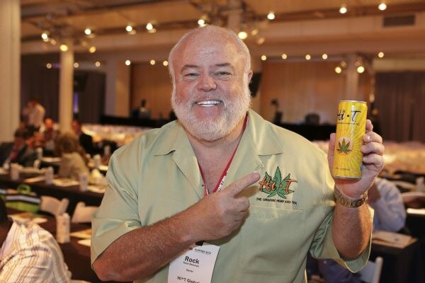 Rock, a smiling bearded man in a green shirt with a Hi-T logo, points to a yellow Hi-T can he holds up, with a conference room of tables behind him.