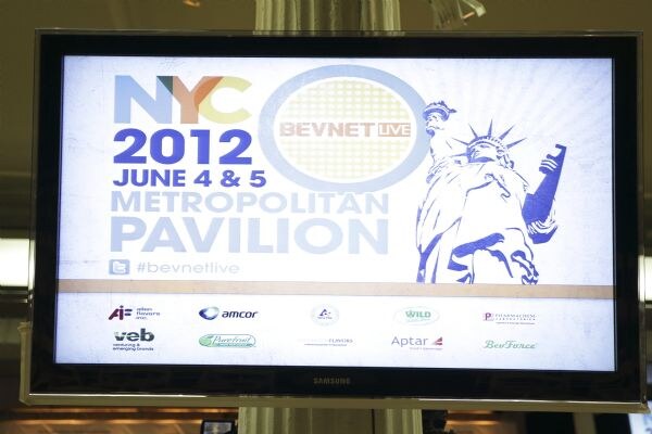 A Samsung monitor shows a BevNET Live graphic reading NYC 2012, June 4 & 5, Metropolitan Pavilion, with a Statue of Liberty illustration, the hashtag #bevnetlive and a row of sponsor logos.