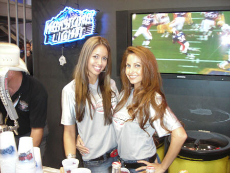 Two smiling women with long hair in gray T-shirts stand beside a neon Mass Comet Light sign, with a TV showing a football game behind them and cups in the foreground.