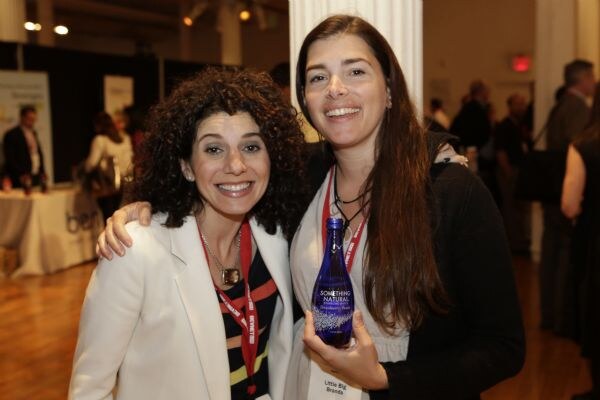 Two smiling women with red lanyards pose close together; the woman on the right holds a blue bottle labeled Something Natural.