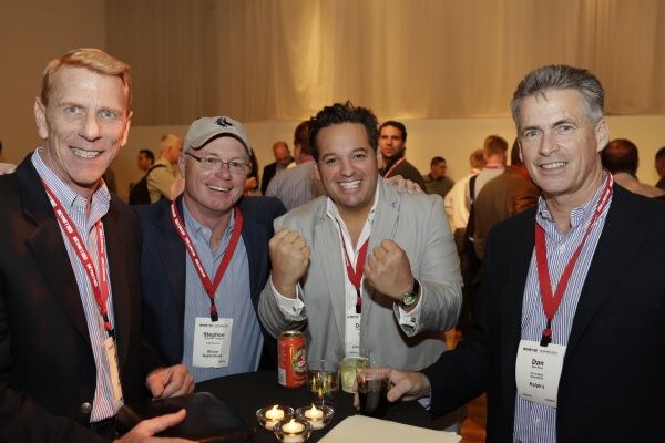 Four smiling men in jackets and red lanyards gather at a table with candles and drinks. Stephen, in a cap, is second from left; Dan is on the right. The man at center raises two fists.
