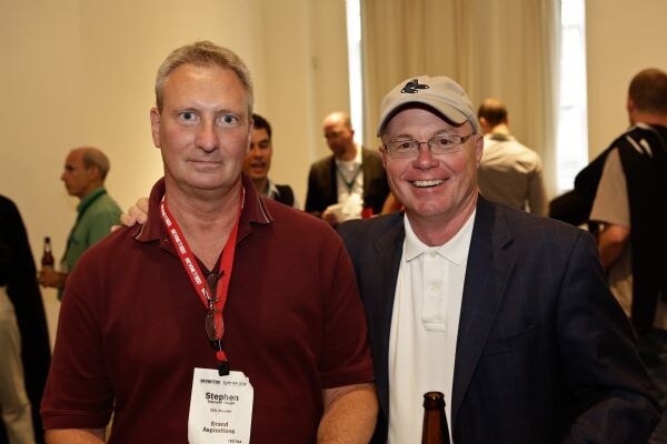 Stephen of Brand Aspirations, on the left in a maroon shirt and red lanyard badge, stands beside a smiling man in a baseball cap and blazer holding a bottle, with other attendees behind them.