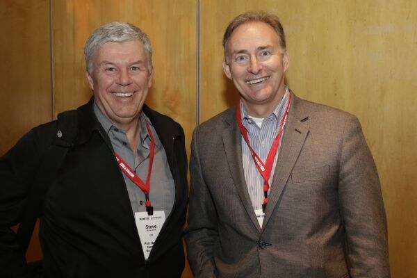 Two smiling men stand together wearing red lanyards. The man on the left, Steve, has gray hair and a dark jacket; the man on the right wears a brown blazer.