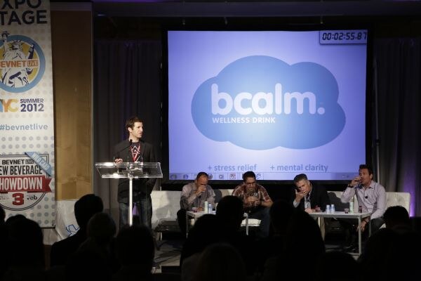 A man speaks at a clear podium on stage beside a screen showing the bcalm wellness drink logo, while four people sit on white couches nearby and the audience watches from the foreground.