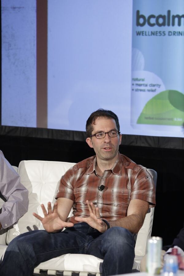 A man with glasses and a plaid short-sleeved shirt gestures with both hands while seated on a white couch, with a bcalm wellness drink can image on the screen behind him.