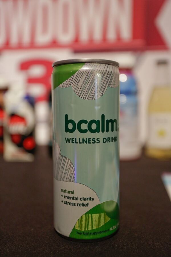 Close-up of a green and white bcalm wellness drink can on a dark table, labeled natural, mental clarity and stress relief, with other blurred bottles and a red Showdown sign behind it.