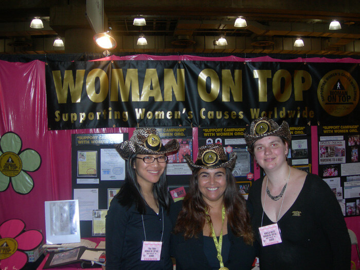 Three women in leopard-print hats and black tops stand smiling in front of a black Woman On Top banner reading Supporting Women's Causes Worldwide, with a pink backdrop and flower decorations.