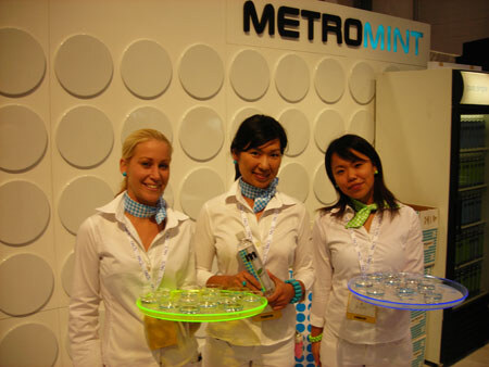 Three smiling women in white shirts and blue-green neck scarves stand in front of a white wall with a Metro Mint sign, two holding colorful lighted trays of small cups.