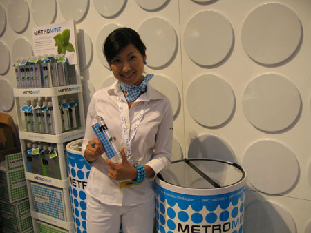 A smiling woman in a white shirt and blue scarf points to a Metromint bottle she holds, beside a Metromint display shelf and a round blue-and-white Metromint cooler bin.