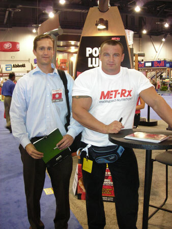 Two men stand in an exhibit hall. One in a white MET-Rx shirt leans on a tall table; the other in a light blue shirt holds a green folder. An Abbott sign is behind.