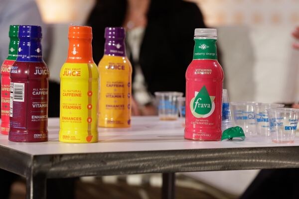 Colorful juice bottles in red, purple, orange and yellow on a table beside a red Frava Cranberry Orange bottle and small plastic sample cups, with blurred people behind.