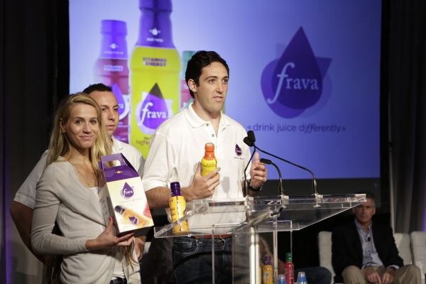 A man in a white polo holds a bottle at a podium while a woman in a gray cardigan holds a Frava carrier box beside him; the screen behind reads Frava, drink juice differently.