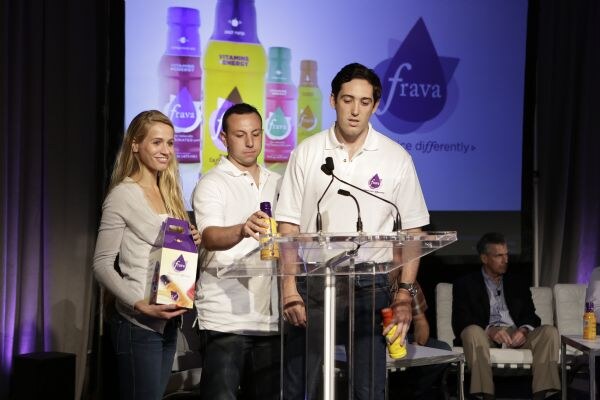 A woman and two men in white polo shirts stand at a clear podium holding Frava juice bottles and a carrier box, with a Frava slide on the screen behind and a seated man at right.