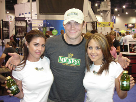A man in a white Mickey's cap and gray Mickey's Fine Malt Liquor shirt stands between two smiling women in white tops, each holding a green Mickey's bottle, in a trade show hall.