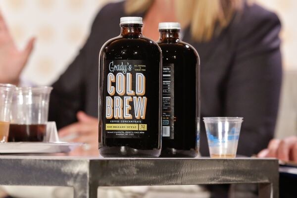 Two dark glass bottles of Grady's Cold Brew coffee concentrate on a metal table, with small plastic cups beside them and a blurred seated person behind.