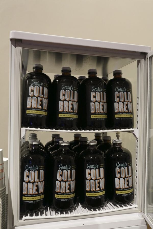 Two shelves of dark glass bottles labeled Grady's Cold Brew New Orleans-Style coffee concentrate stacked inside a small white display refrigerator, with paper cups at the left edge.