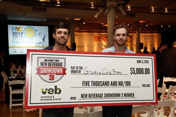 Two men stand holding a large check for $5,000 payable to Grady's Cold Brew, labeled New Beverage Showdown 3 Winner with the veb logo, in an event space with chairs and a projection screen.