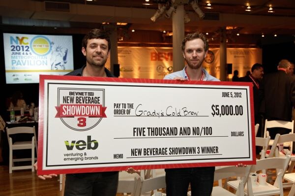 Two men hold a large novelty check for $5,000 made out to Grady's Cold Brew, marked New Beverage Showdown 3 Winner with a veb logo, in an event hall with a NYC 2012 screen behind them.