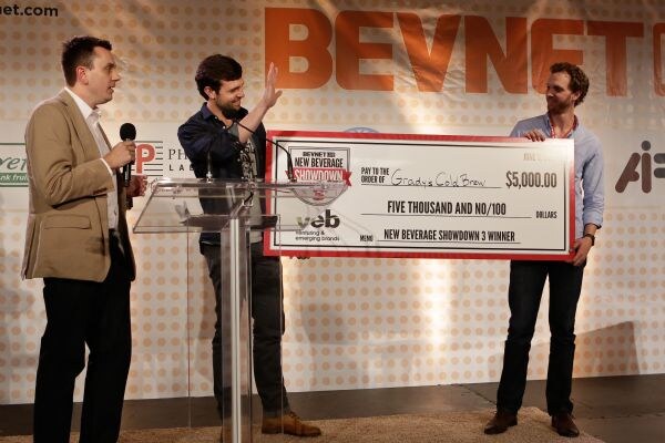 Three men on stage: one holds a microphone, one waves at a lectern, and one holds a large $5,000 check made out to Grady's Cold Brew for the New Beverage Showdown 3 winner.