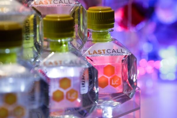 Close-up of small angular clear Last Call Productivity Beverage bottles with olive-green caps and orange hexagon Lc+ logos, against a blurred purple and pink background.