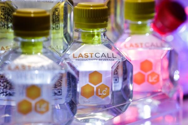 Close-up of a row of clear Last Call Productivity Beverage bottles with green caps and orange hexagon Lc+ logos, with pink and purple light in the background.