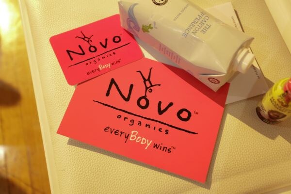 Two pink Novo Organics cards reading everyBody wins lie on a white leather surface next to a carton of water and a small yellow bottle.