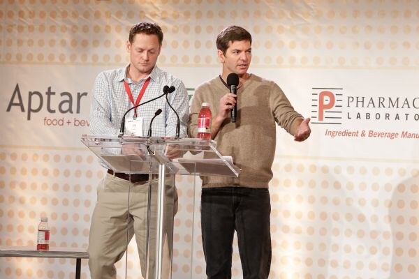 Two men stand on stage at a clear podium in front of a banner for Aptar and Pharmachem Laboratories. One holds a microphone and gestures with his arm; a red drink bottle sits on the podium.