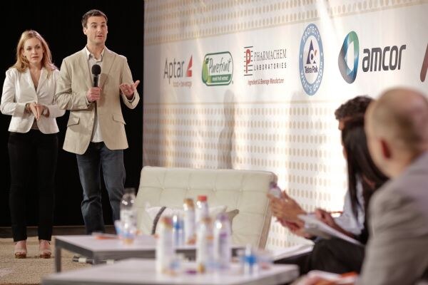 A woman in white and a man in a tan blazer holding a microphone stand on stage before a banner with Aptar, Purefruit, Pharmachem Laboratories, Tetra Pak and Amcor logos; judges sit in the foreground.