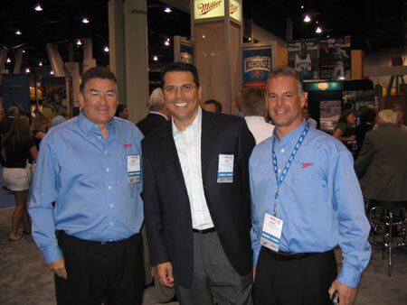 Three smiling men with name badges pose together in a trade-show hall, two in light blue button-up shirts and one in a dark blazer, with Miller and Icehouse signs behind them.
