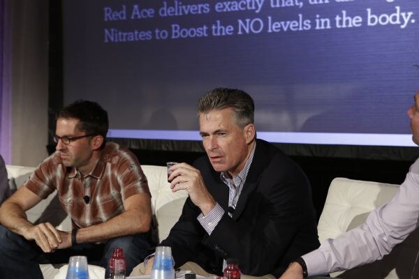 A man in a dark blazer holds a small cup while speaking, seated beside a man in a plaid shirt and glasses, with bottles and cups on a table and a Red Ace slide behind.