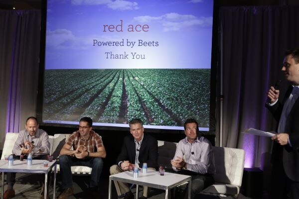 Four men sit on white couches beside low tables with cups, facing a screen reading red ace, Powered by Beets, Thank You. A man at right holds a microphone and papers.