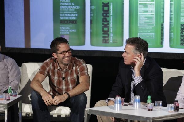 A smiling man in a plaid shirt and a man in a dark blazer, hand at his ear, sit on white couches in front of a screen of green RuckPack bottles, with small cups and bottles on a table.