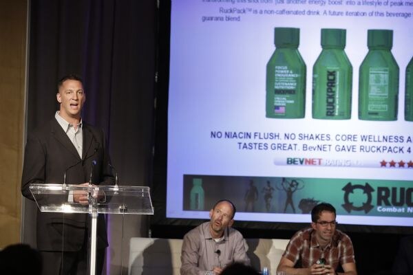 A man in a dark jacket speaks at a clear podium while a screen behind him shows RuckPack bottles and a BevNET rating. Two men sit on a white couch below the screen.