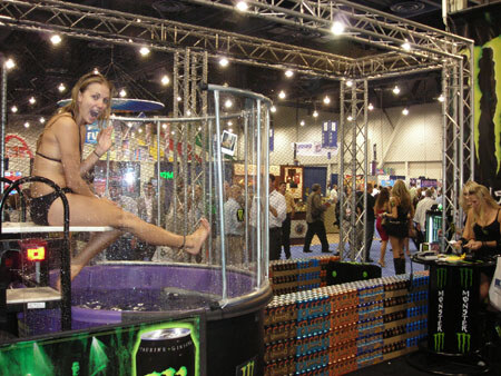A woman in a bikini sits on the edge of a glass dunk tank with her legs hanging over. Stacked cases and Monster Energy branding surround the tank in a busy exhibit hall.