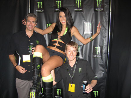 A woman in a black outfit and tall black boots sits on a man's raised leg between two men in black Monster Energy shirts, holding cans, in front of a Monster Energy logo backdrop.