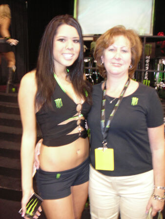 A woman in a black Monster Energy top and shorts stands beside a woman with a yellow badge on a lanyard wearing a dark Monster Energy shirt, posing together at a trade show.