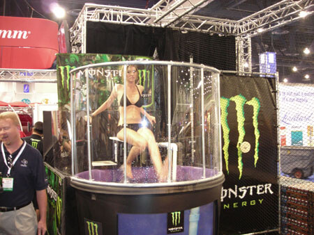 A woman in a bikini sits on a seat above water in a clear Monster Energy dunk tank with Monster Energy banners, while a man in a dark polo and lanyard stands at left.