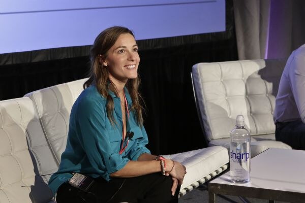 A smiling woman in a teal blouse sits on a white tufted couch on stage with a microphone clipped on. A bottle of Sharp Water stands on the table in front of her.