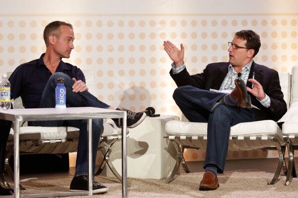 Two men sit in white leather chairs on stage. One gestures with a raised hand while the other listens, and a Zico bottle stands on the table between them.