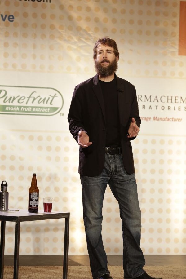 A bearded man in a dark blazer and jeans gestures while speaking on stage. A bottle of Arrogant Bastard Ale and a glass of red liquid sit on a small table beside him.