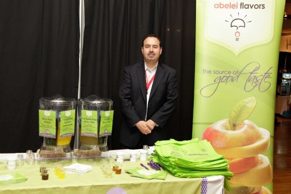 A man in a dark suit with a red lanyard stands behind an Abelei Flavors booth table with two beverage dispensers, sample cups and green bags, beside a green Abelei Flavors banner.