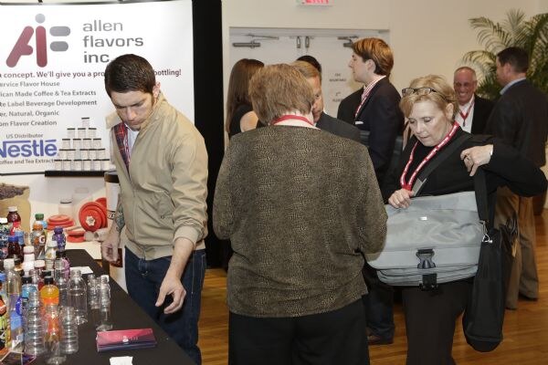 Attendees with lanyards gather at the Allen Flavors booth, where bottles and sample vials sit on a dark table. The banner also shows the Nestle name.