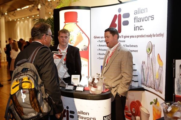 Two men in jackets, one wearing a backpack, talk with a man in a tan blazer at the Allen Flavors booth, in front of a banner reading allen flavors inc. and a lit bottle graphic.