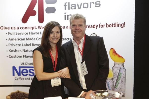 A smiling woman with long dark hair and a smiling man in a dark jacket, both wearing lanyards and badges, pose together in front of an Allen Flavors banner.
