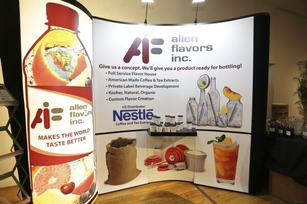 Empty Allen Flavors booth with a curved backdrop reading allen flavors inc., a lit bottle graphic reading Makes the world taste better, a Nestle logo and a shelf of sample jars.