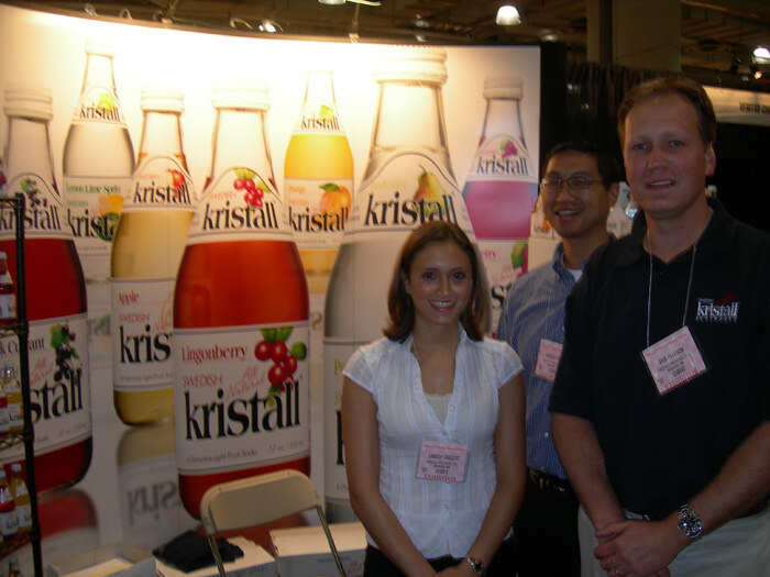 Two men and a woman pose at a booth with large Kristall Swedish soda bottle graphics, including Lingonberry and Apple flavors, behind them. Each wears a name badge.