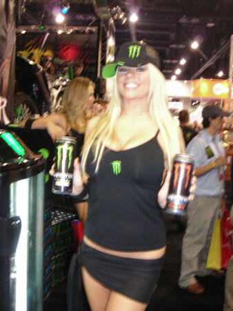 A smiling woman with long blonde hair wears a black Monster Energy cap and top and holds a Monster Energy can in each hand at a trade show booth.