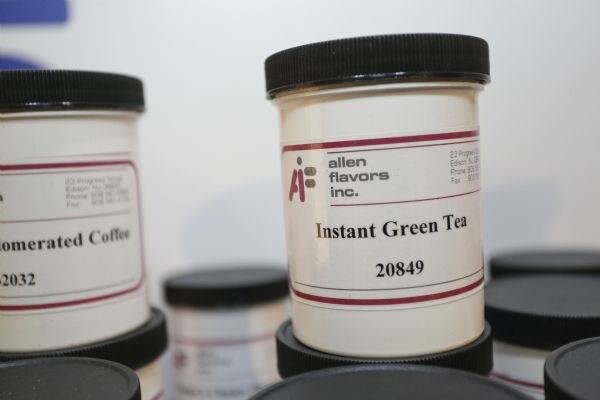 Close view of white sample jars with black lids and Allen Flavors Inc. labels, including one labeled Instant Green Tea 20849 and another partly visible labeled Coffee.